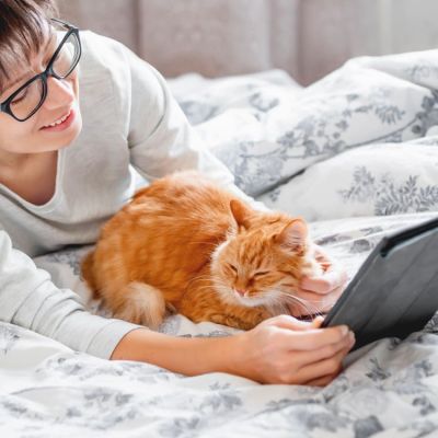 Cute ginger cat and woman in glasses are lying in bed. Woman is